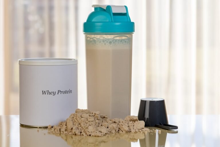 Bote de proteína whey junto a un vaso mezclador con batido, un cacito y proteína en polvo sobre la mesa